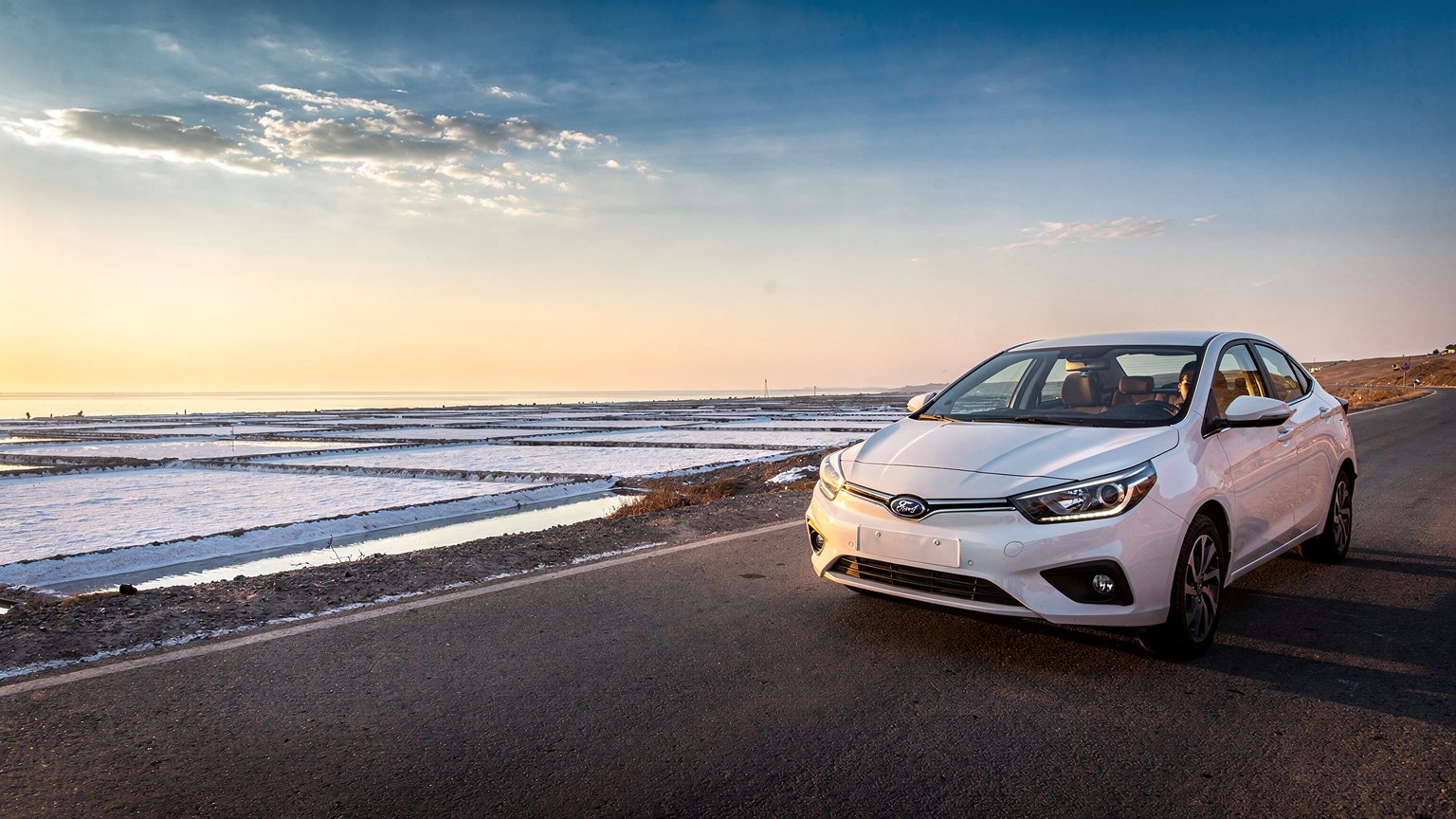 White sedan beside salt pans on a coastal road (illustrative image)